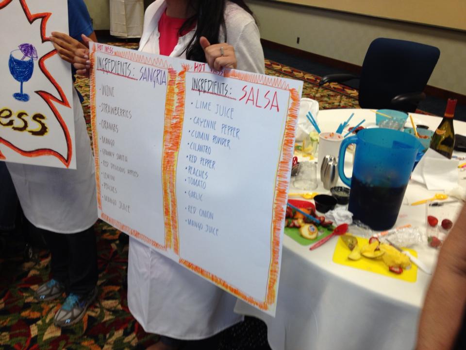 A team member holds up ingredient lists for Sangria and Salsa at the Salsa Showdown team building event, with colorful ingredients and materials on the table.