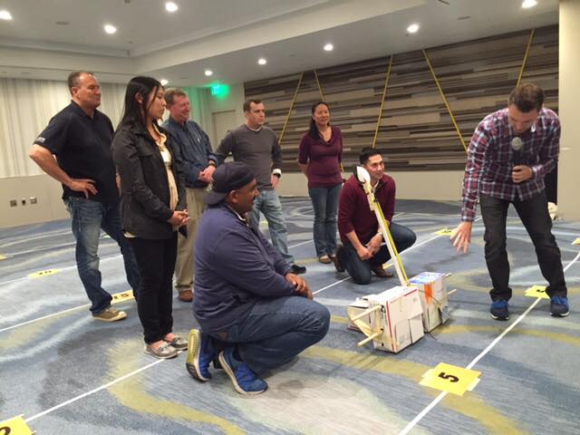 A group of participants indoors discussing their catapult design as part of the Catapult to Success team building activity, focusing on teamwork, strategy, and collaborative problem-solving in a competitive setting.