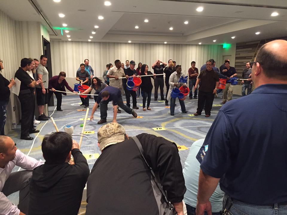 Participants in an indoor setting engaging in a competitive team building activity with catapults, buckets, and various objects, demonstrating teamwork, strategy, and coordination during the Catapult to Success event.