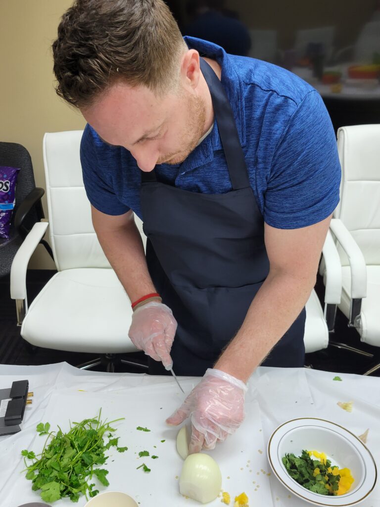 A man in a blue shirt and black apron is carefully chopping an onion at a table during a team building chili cook-off event. He is wearing disposable gloves, and there are herbs, chopped vegetables, and a small bowl with ingredients on the table in front of him.