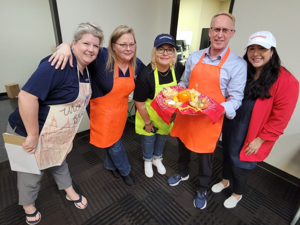 A group of five people smiling and posing, holding a tray of food. They are participating in a chili cook-off team building activity. Three of the participants wear bright aprons, one in orange and one in lime green. The man in the center is holding a red tray with an arrangement of colorful vegetables and food items.