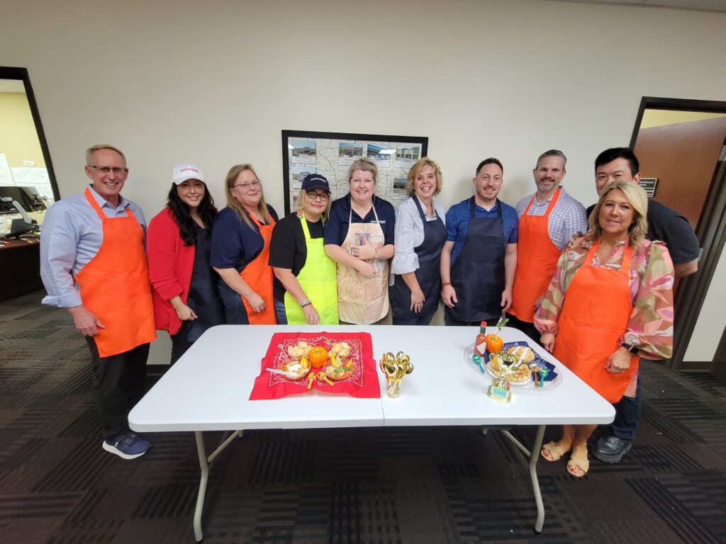 A group of individuals stands together, smiling, behind a table at a team building chili cook-off event. Some are wearing brightly colored aprons in shades of orange, blue, and green. The table in front of them displays two creatively presented chili dishes, along with small golden trophies.