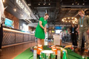 A participant plays on a creatively designed mini golf course made from canned goods and food packages during a 'Build Your Own Mini Golf Course' team building event. The event takes place in a lively venue, combining fun, teamwork, and charity, with the food items used for the course being donated to a local food pantry afterward.