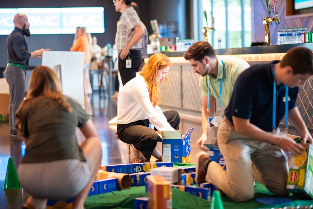 Participants collaborate to build a mini golf course using canned goods and packaged food items during a 'Build Your Own Mini Golf Course' team building event. The activity encourages teamwork and creativity, with the food items being donated to a local food pantry after the event, combining fun with a charitable cause.