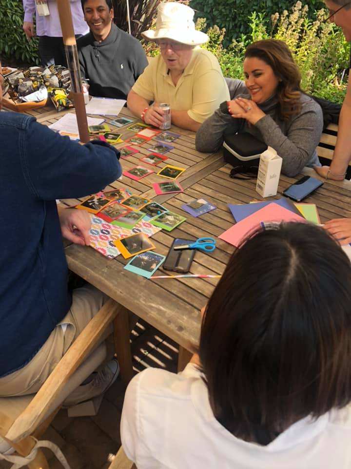 A group of team members gathered around a wooden table outdoors, collaborating on a photo collage as part of a Polaroid scavenger hunt. The table is filled with colorful paper, Polaroid photos, scissors, and crafting materials as participants engage in friendly conversation and creative teamwork, enjoying a relaxing and productive moment in a sunny garden setting.