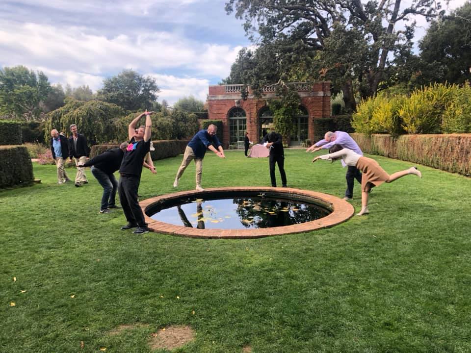 A group of team members engage in a playful pose around a small circular pond in a lush garden during an outdoor team building event.