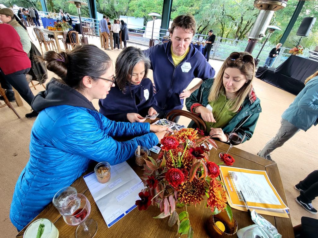 Four team members engage in a fun Polaroid scavenger hunt, reviewing instant photos taken during an outdoor team building event. The group gathers around a table with vibrant fall-themed flower arrangements and drinks.