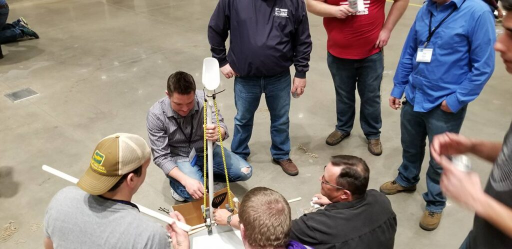 A group of participants indoors gathered around a table, actively assembling a catapult as part of the Catapult to Success team building activity, focusing on collaboration, innovation, and hands-on problem-solving.