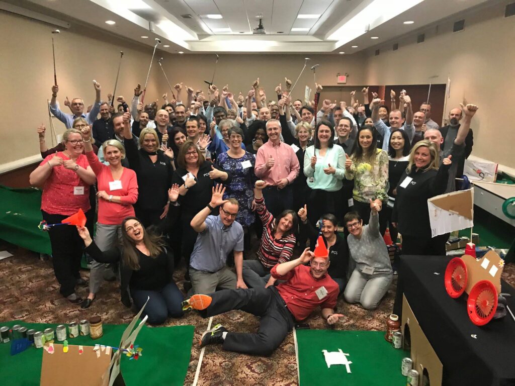 A large group of participants celebrating together after a successful 'Build Your Own Mini Golf Course' team building event. The group poses with their hands raised and mini golf clubs in hand, surrounded by creative mini golf holes made from canned goods and other non-perishable food items.