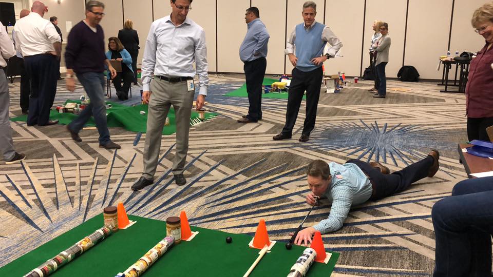 A participant lying on the floor carefully lines up a shot during a 'Build Your Own Mini Golf Course' team building event. The mini golf course is constructed from canned goods and other food items, promoting both fun teamwork and charity, with the food being donated to a local pantry afterward.