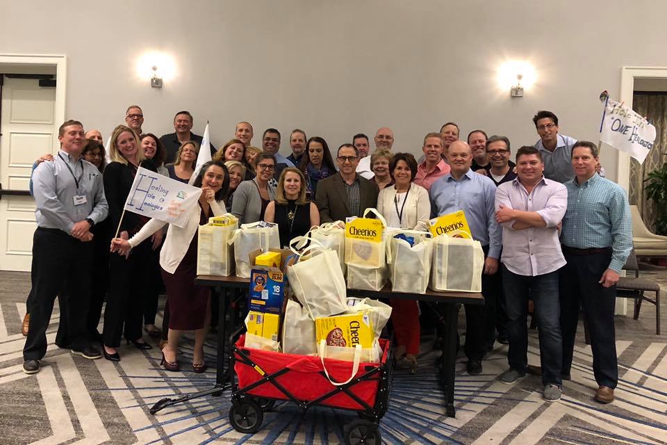 A group of participants proudly stands behind a collection of bags filled with groceries after a successful 'Build Your Own Mini Golf Course' team building event. The event encourages teamwork and charitable giving, with the food items being donated to a local food pantry to support families in need.