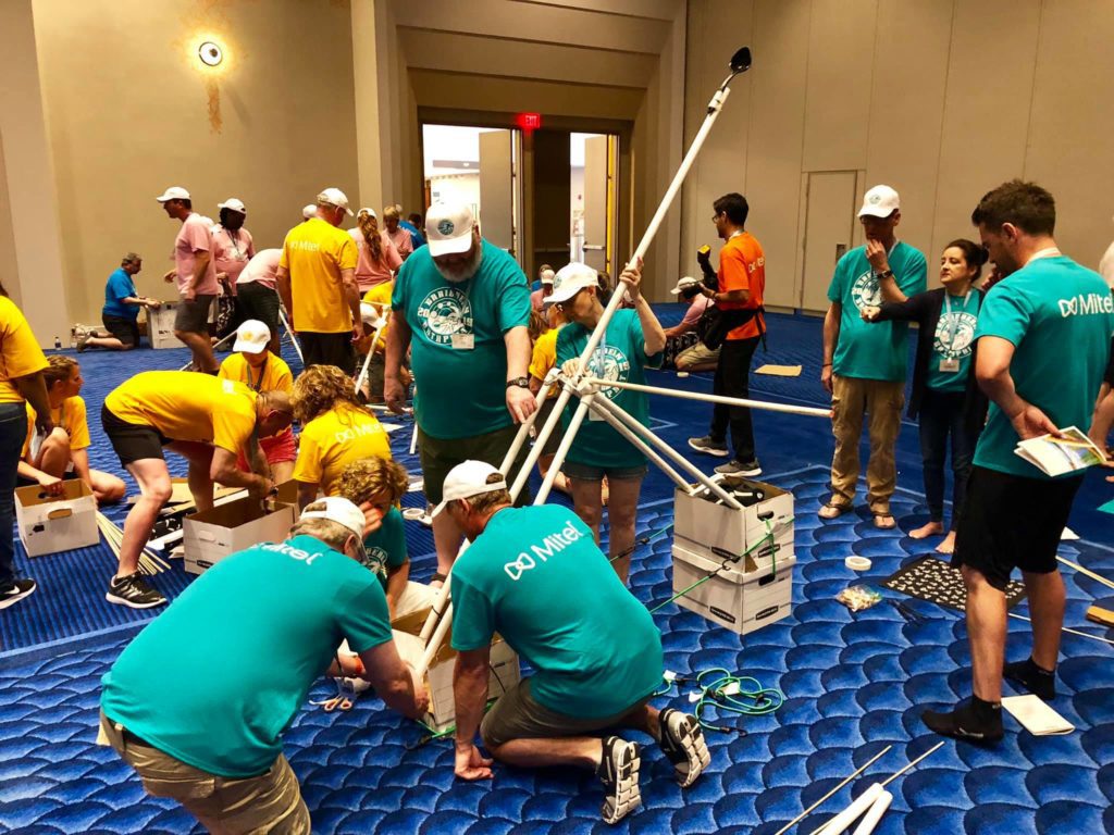 Multiple teams in brightly colored shirts collaborating on building catapults indoors during the Catapult to Success team building activity, showcasing teamwork, creativity, and hands-on construction skills in a lively group setting.