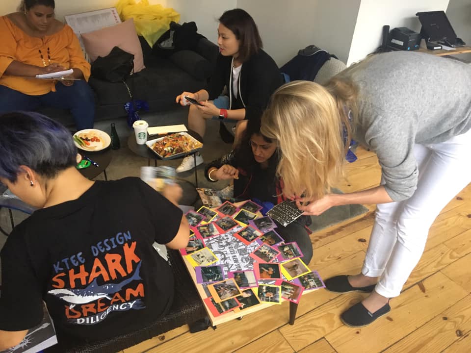 A dynamic scene of team members assembling a collage on the floor, using Polaroid photos from a team building scavenger hunt. Individuals focus on arranging the photos, and enjoying light snacks. The activity fosters teamwork and creativity.