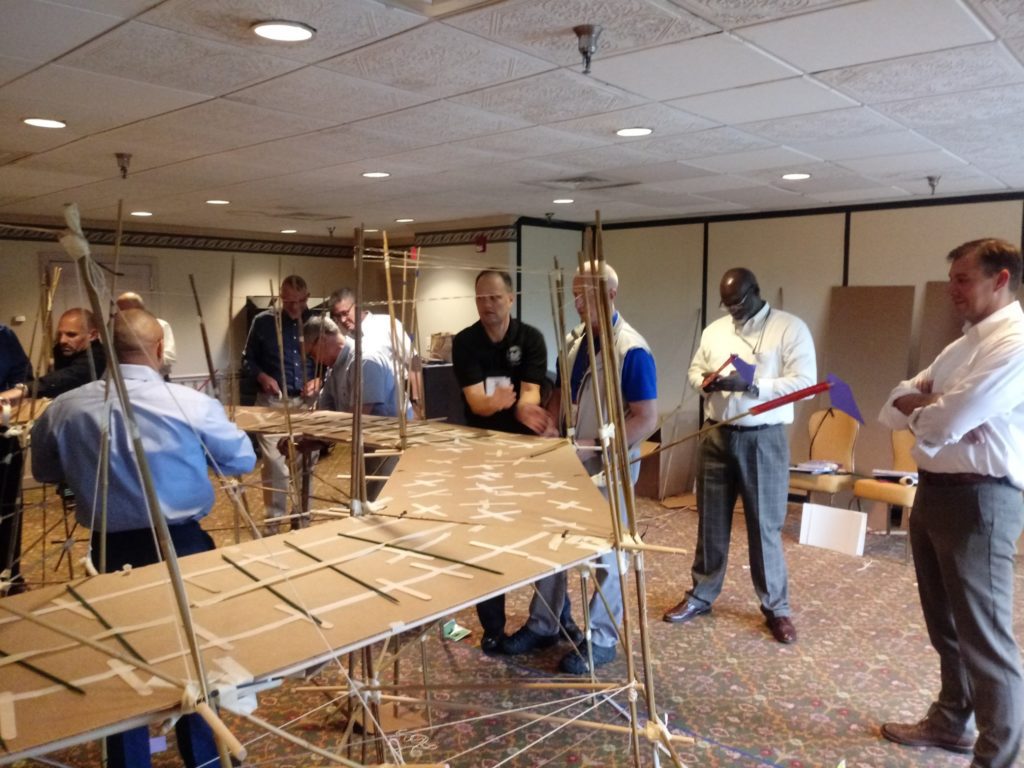 Participants in the Bridging the Divide team building activity gather around a large, collaboratively built cardboard bridge, assessing its structure and stability.