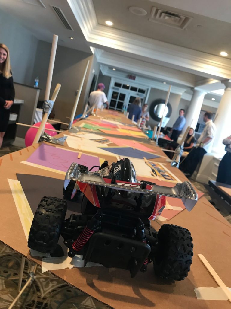 A remote-controlled car prepares to cross a large cardboard bridge designed by participants during the Bridging the Divide team building event.