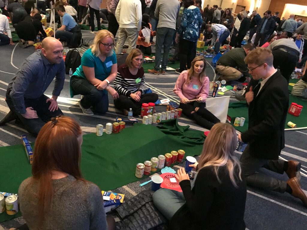 A group of participants collaborating on building a mini golf course using canned goods and non-perishable food items during a 'Build Your Own Mini Golf Course' team building event.