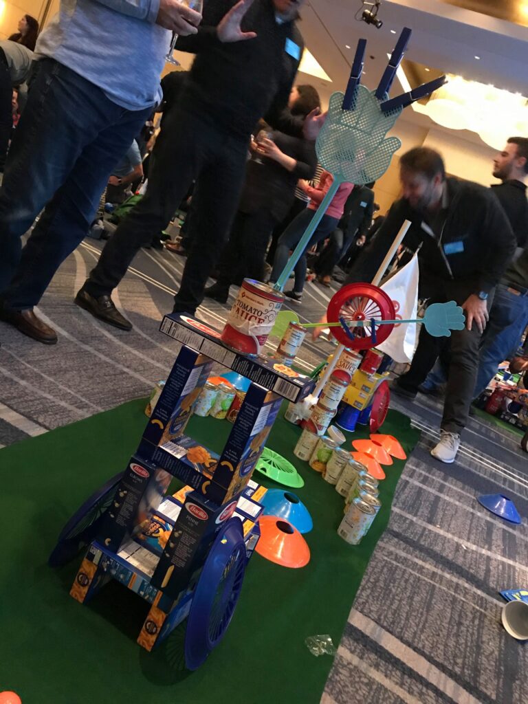 A creatively designed mini golf hole built from food cans, pasta boxes, and other non-perishable items during a 'Build Your Own Mini Golf Course' team building event. The course features a playful structure with a toy wheel and hand decorations.