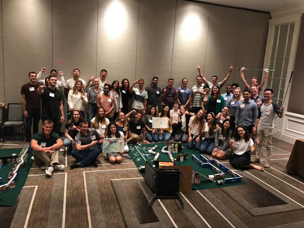 A large group of participants celebrates together after a successful 'Build Your Own Mini Golf Course' team building event. The group poses with their hands raised, surrounded by mini golf courses creatively constructed from canned goods and other non-perishable items. The event fosters teamwork and contributes to charity, with the food items being donated to a local food pantry.
