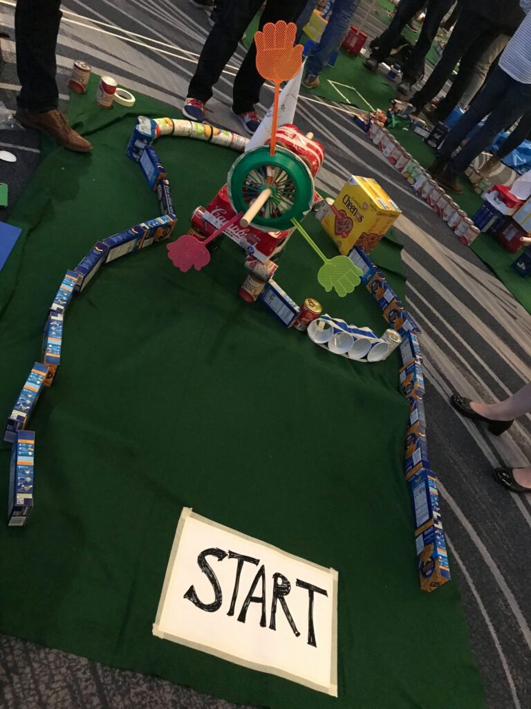 A creative mini golf course built from food packages, including cereal boxes, cans, and snack items, with a 'Start' sign marking the beginning of the hole. Participants designed the course as part of a 'Build Your Own Mini Golf Course' team building event, combining fun, creativity, and charity. The food items used are later donated to a local pantry.