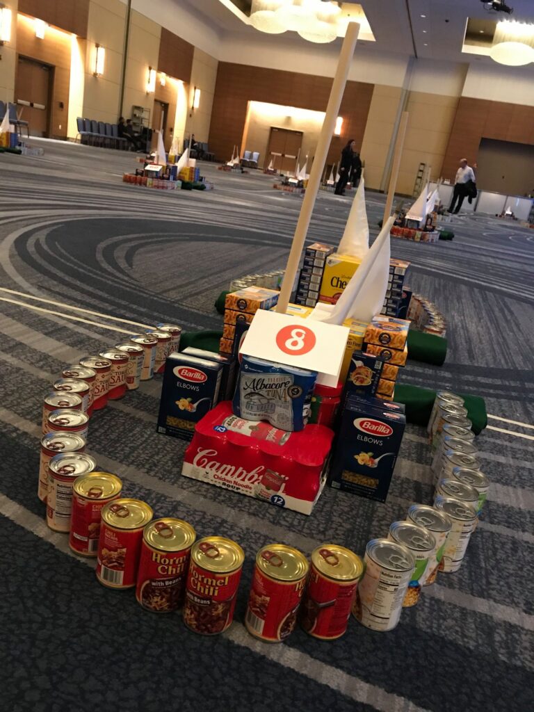 A mini golf course hole constructed from food cans, cereal boxes, and other non-perishable items during a 'Build Your Own Mini Golf Course' team building event.