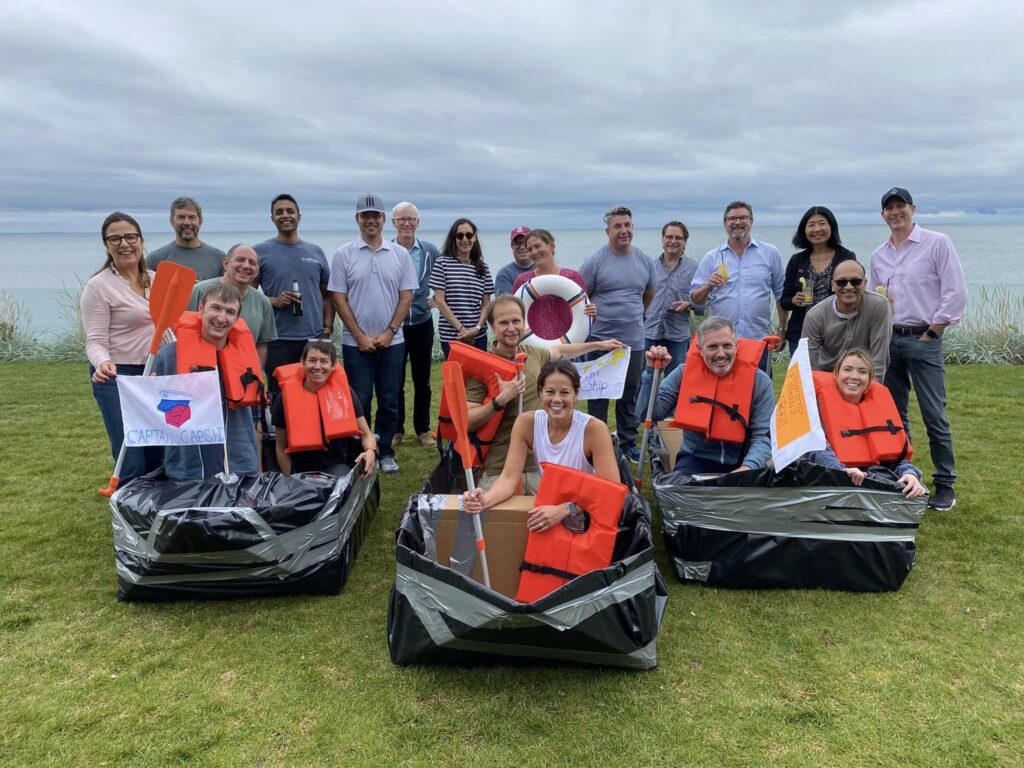 Team posing with their decorated cardboard boats and flags during the Build-A-Boat team building challenge near the water.