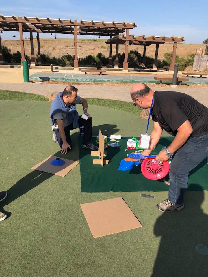Two participants engaged in a team building 'Build Your Own Mini Golf Course' event, working on constructing a mini golf hole using cardboard, a fan, and other creative materials on a mat laid out on a grassy area outdoors. This activity encourages teamwork and innovation while using everyday objects to create a golf course..