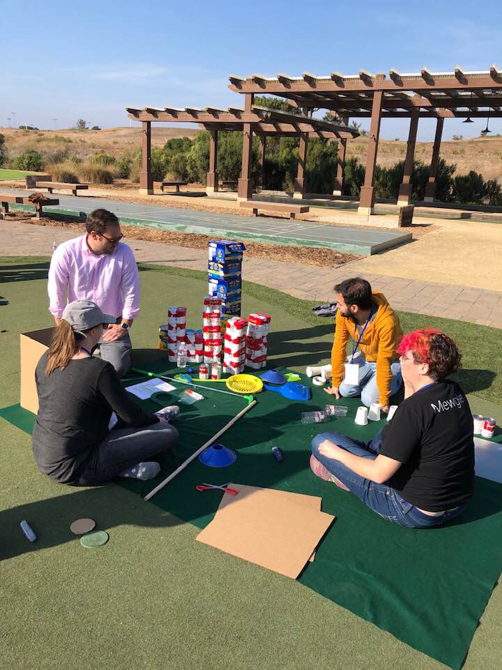 Group of participants sitting on a mat outdoors, collaborating to build a creative mini golf hole using food cans, boxes, and various supplies during a 'Build Your Own Mini Golf Course' team building event.