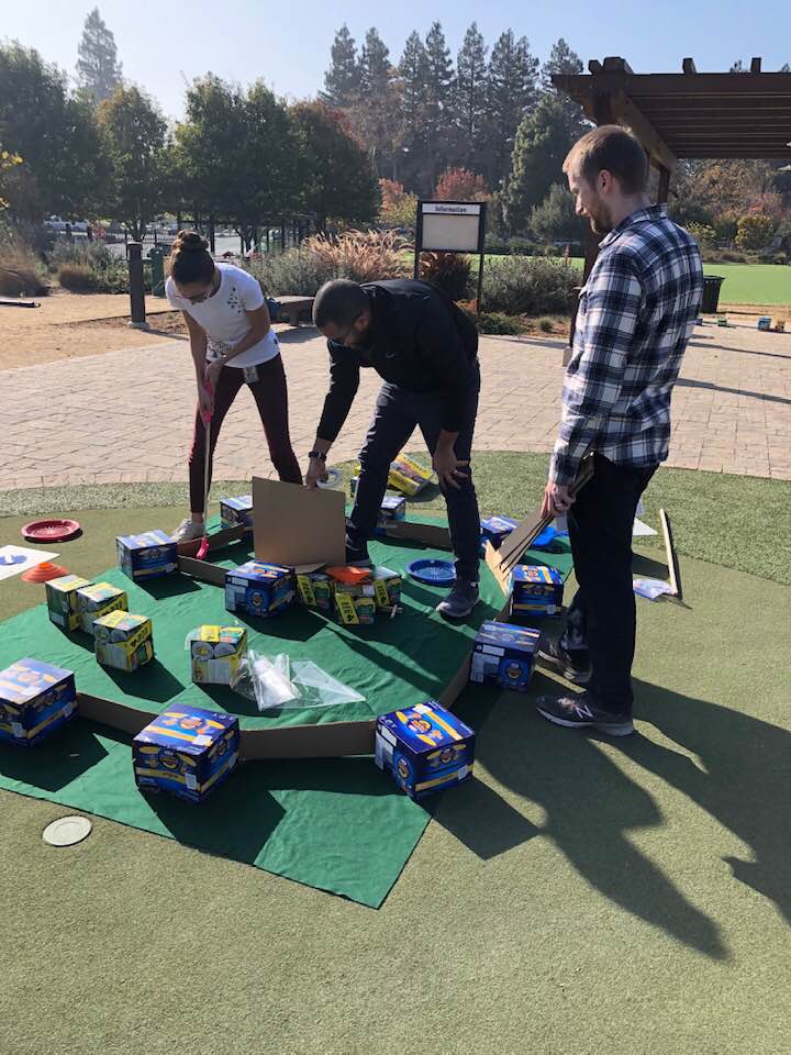 Participants collaborate to build a mini golf hole using boxes of food and other non-perishable items during an outdoor 'Build Your Own Mini Golf Course' team building event. The activity combines creativity, teamwork, and charity, with the food items being donated to a local food pantry afterward.