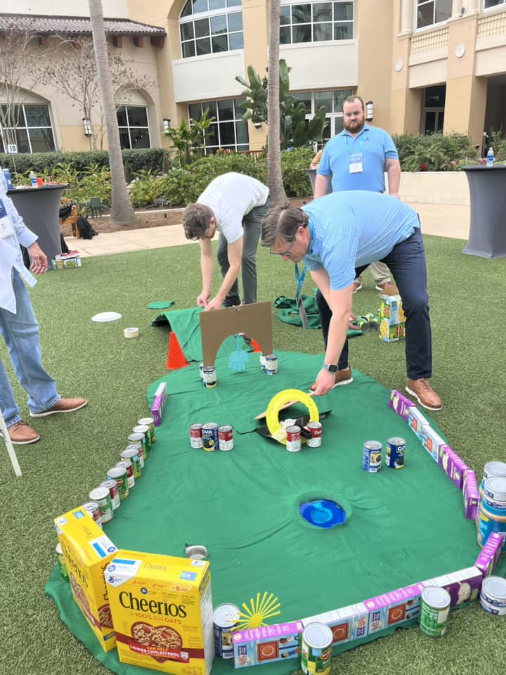 Participants setting up a creative mini golf course using canned goods, cereal boxes, and cardboard during a 'Build Your Own Mini Golf Course' team building event.