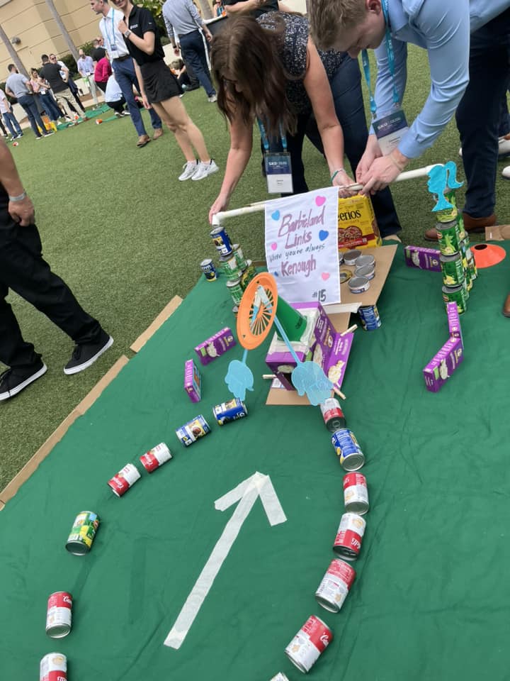 Participants assemble a creative mini golf hole using canned goods and food packages during a 'Build Your Own Mini Golf Course' team building event. The course features a sign labeled 'Birdland Links' and is designed with fun, colorful elements.
