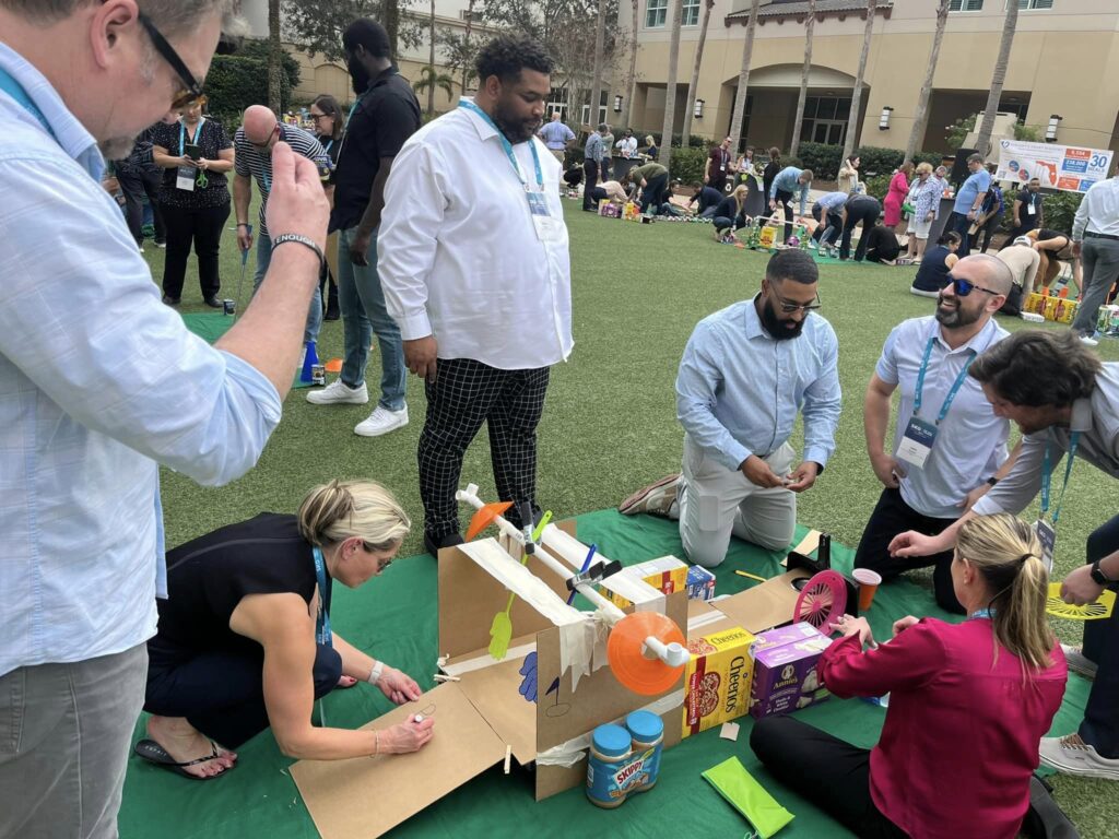 A team of participants collaboratively building a creative mini golf hole using cardboard, canned goods, and food packages during a 'Build Your Own Mini Golf Course' team building event.