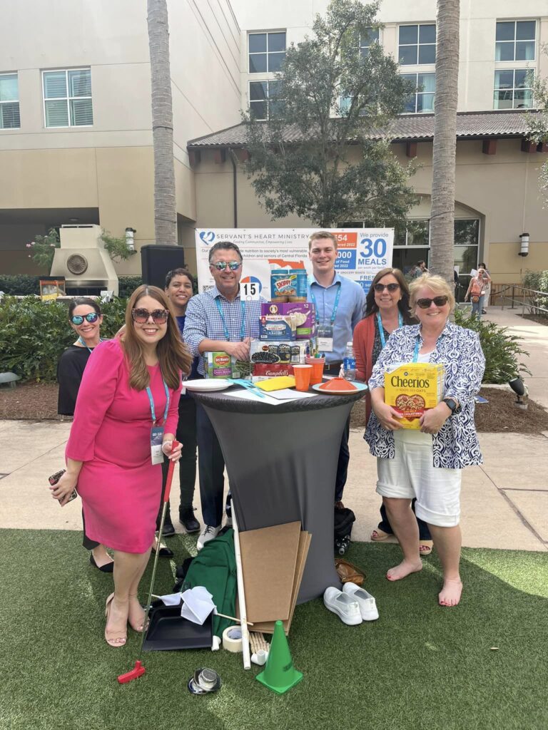 A group of participants posing with canned goods and food packages during a 'Build Your Own Mini Golf Course' team building event. The team stands outdoors, displaying their collection of food items that will be donated to a local charity, combining creativity, teamwork, and a meaningful cause.