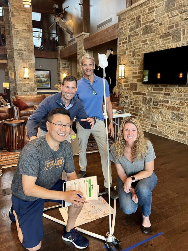 A group of four participants smiling and posing indoors with a constructed catapult during the Catapult to Success team building activity, emphasizing teamwork, innovation, and collaborative problem-solving.