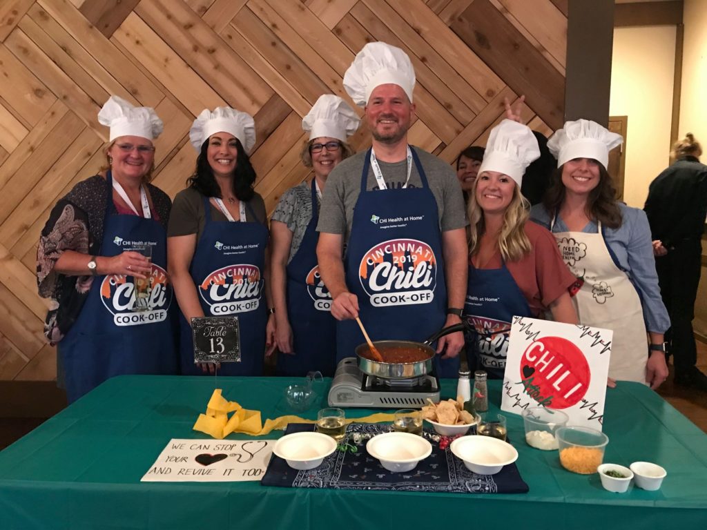 A group of seven people stand behind a table, smiling and wearing chef hats and aprons for a Chili Cook-Off event. They are preparing a chili dish with a pot on a portable stove in front of them. Various ingredients and garnishes are laid out on the table. A sign reads 'We can stop your heart and revive it too!' indicating a playful theme. The group is enjoying a team building culinary event.