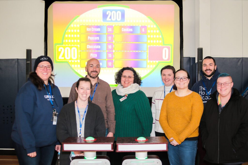 A group of eight participants smiling and standing together in front of a game show setup with a brightly colored game board in the background during the Corporate Feud game.