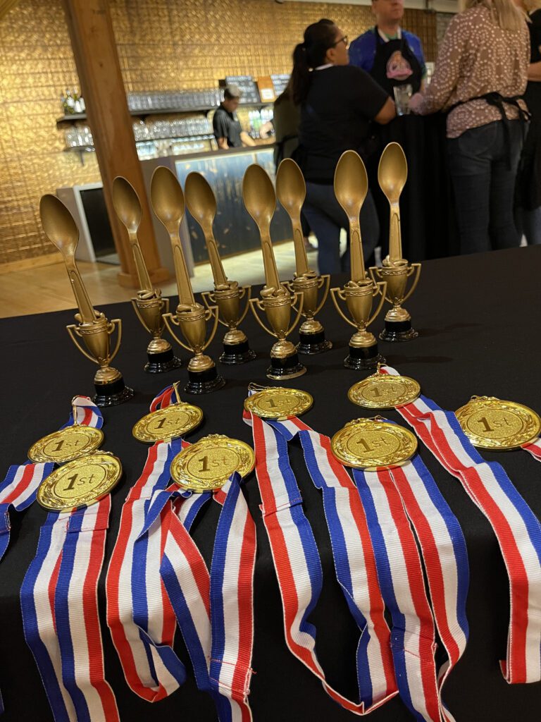 A table displays several gold trophies in the shape of large spoons and medals with '1st' on them. The medals are attached to red, white, and blue ribbons. The trophies and medals are for the culinary team building winners.