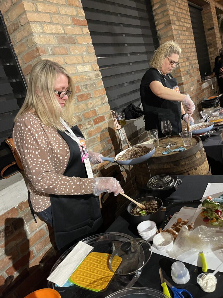 Two women participating in a team building chili cook-off. Both are focused on preparing food at their respective stations. The woman on the left is stirring ingredients in a bowl, wearing a brown polka-dotted cardigan over her apron. The woman on the right is cooking something on a skillet while wearing a black shirt and apron.