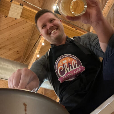 A man is smiling as he participates in a chili cook-off. He is wearing a black apron with the 'Chili Cook-Off' logo prominently displayed. The wooden ceiling and string lights in the background create a cozy atmosphere. The man is holding a glass of beer in one hand and appears to be stirring a pot with the other, fully engaged in the cooking activity.