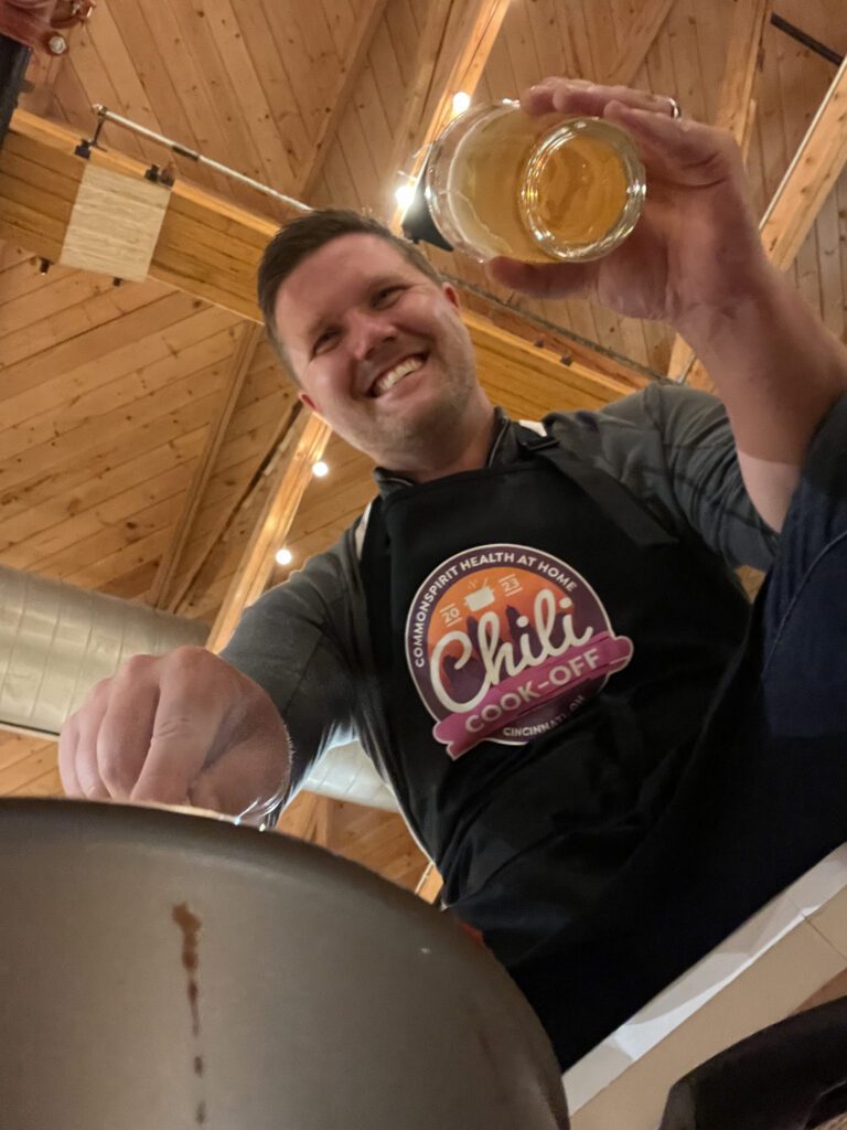 A man is smiling as he participates in a chili cook-off. He is wearing a black apron with the 'Chili Cook-Off' logo prominently displayed. The wooden ceiling and string lights in the background create a cozy atmosphere. The man is holding a glass of beer in one hand and appears to be stirring a pot with the other, fully engaged in the cooking activity.