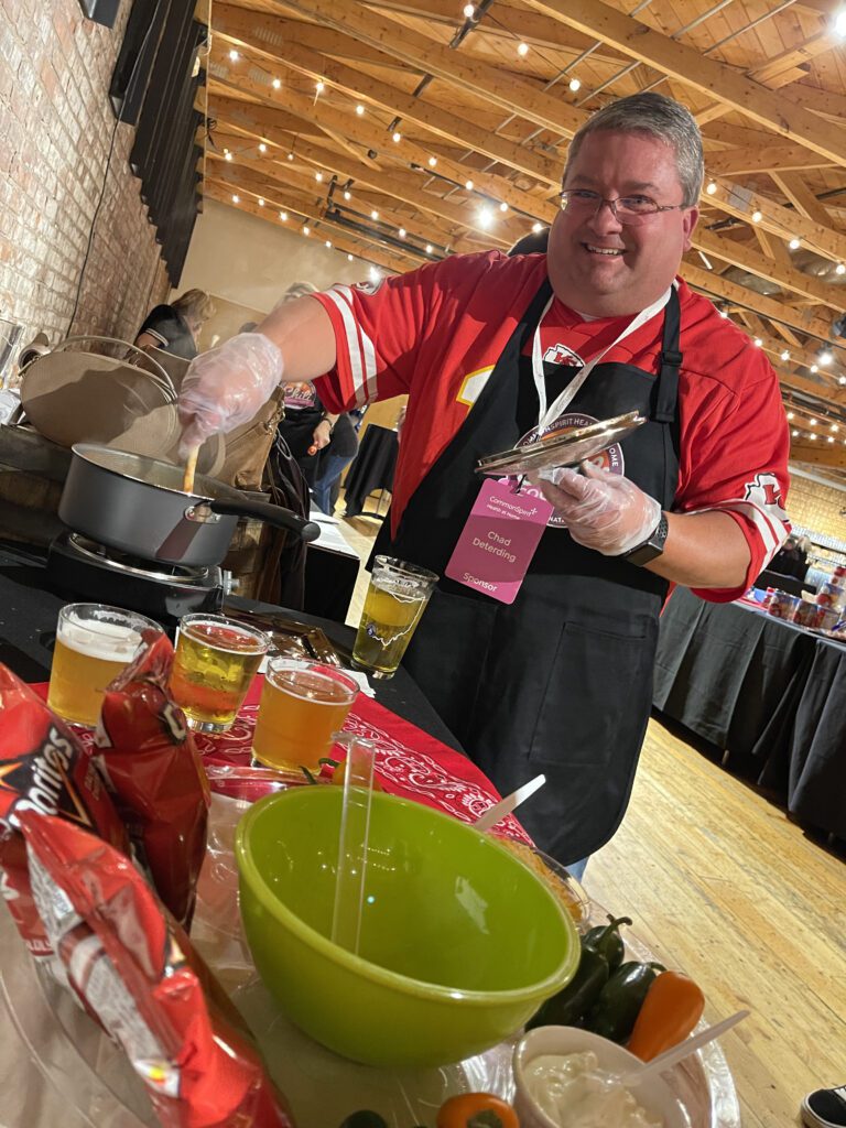 A man wearing a red sports jersey and a black apron is smiling while stirring a pot on a stove during a culinary team building event. He holds a set of tongs in one hand and is wearing plastic gloves. In front of him are cups of beer, a bag of Doritos chips, and a green bowl.
