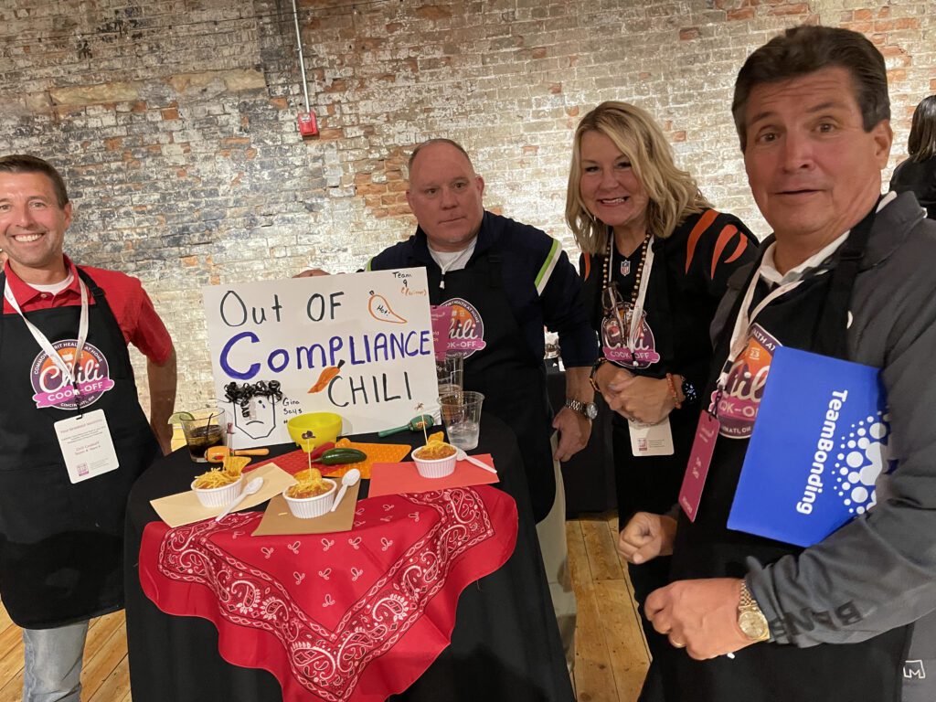 Four people standing around a small round table with a red bandana tablecloth. The group is participating in a chili cook-off team building event. The table features a sign that reads 'Out of Compliance Chili' with some illustrations, along with bowls of chili. The group is smiling and wearing aprons with chili cook-off branding.