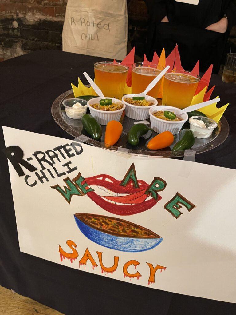 A creatively arranged display featuring small cups of chili garnished with shredded cheese and jalapeños, paired with a dollop of sour cream on the side. The setup includes three glasses of beer placed behind the chili cups, with plastic spoons inserted into each. The presentation is adorned with paper flames, giving a fiery theme to the display. In front, there is a hand-drawn sign that reads 'R-Rated Chili, We Are' alongside a bold illustration of a smiling mouth and a bowl of chili. Several real jalapeño and yellow peppers are placed around the setup.