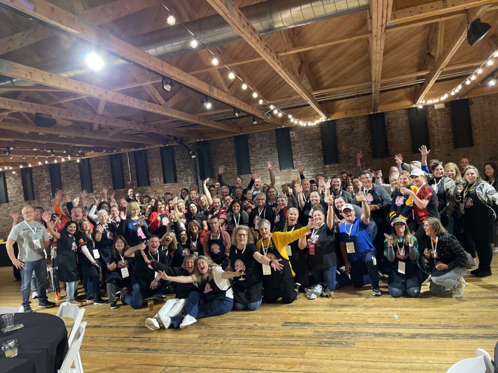 A large group of people posing together in a celebratory manner at an indoor chili cook-off team building. The participants are smiling, waving, and cheering while wearing aprons with chili cook-off branding.