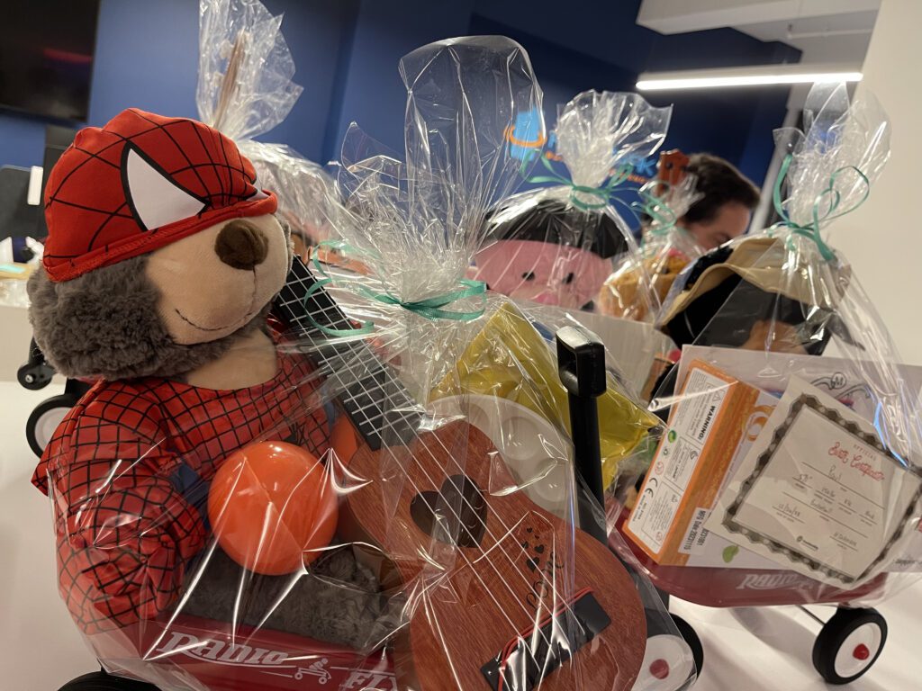 A close-up image of a red Radio Flyer wagon filled with a gift set for charity. The wagon includes a stuffed teddy bear dressed in a superhero outfit, a small ukulele, a maraca, and various art supplies. The entire wagon is wrapped in clear gift wrap, ready for donation at the Toys for Tykes ‘Arts & Music’ team building event.