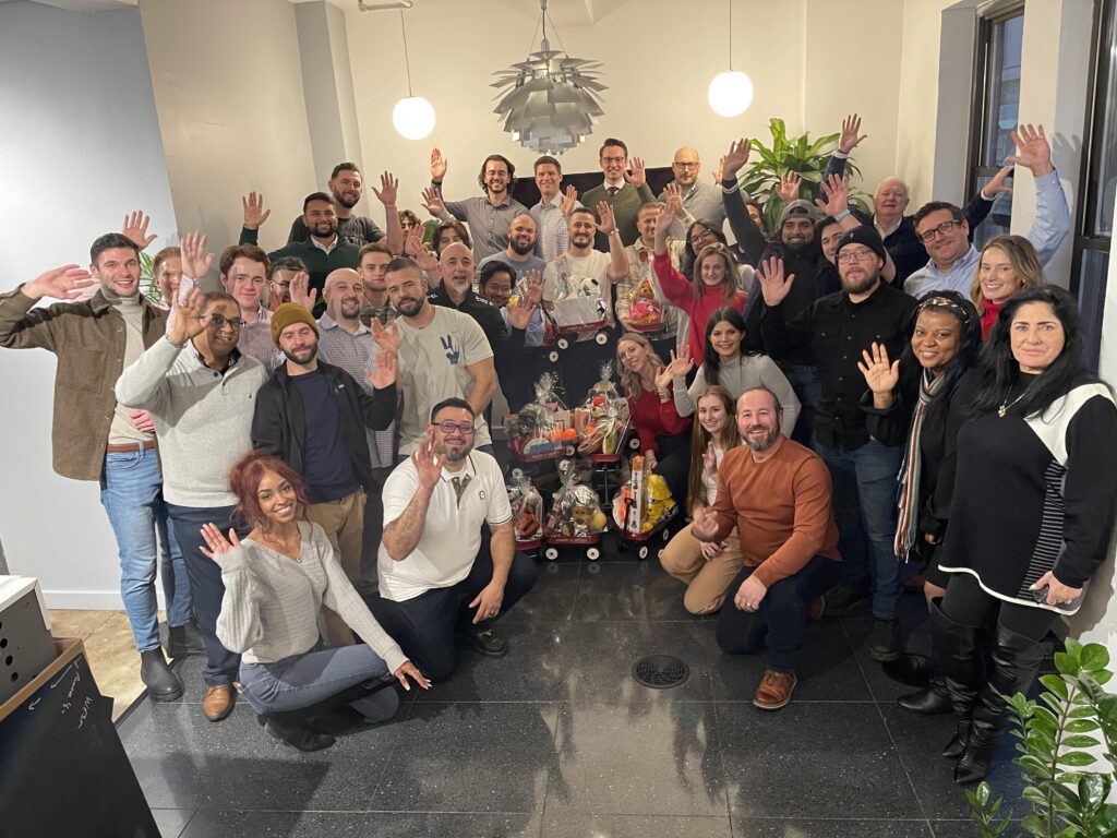 A large group of people smiling and waving for a group photo. They are gathered around a collection of decorated toys and gifts, assembled during a CSR team building program.