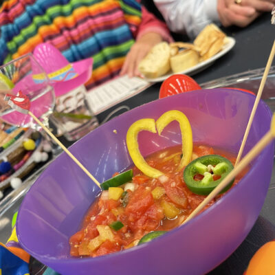 A colorful bowl of salsa garnished with heart-shaped yellow peppers and jalapeños at a Salsa Showdown team building event, with participants wearing vibrant striped outfits.