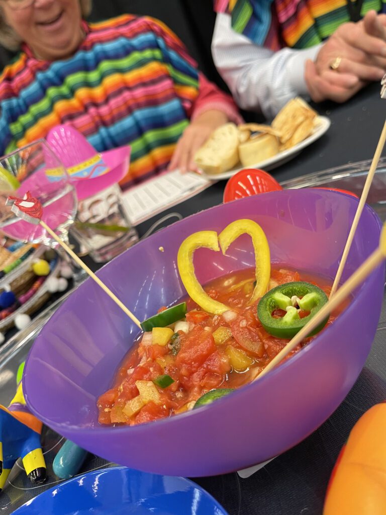A colorful bowl of salsa garnished with heart-shaped yellow peppers and jalapeños at a Salsa Showdown team building event, with participants wearing vibrant striped outfits.