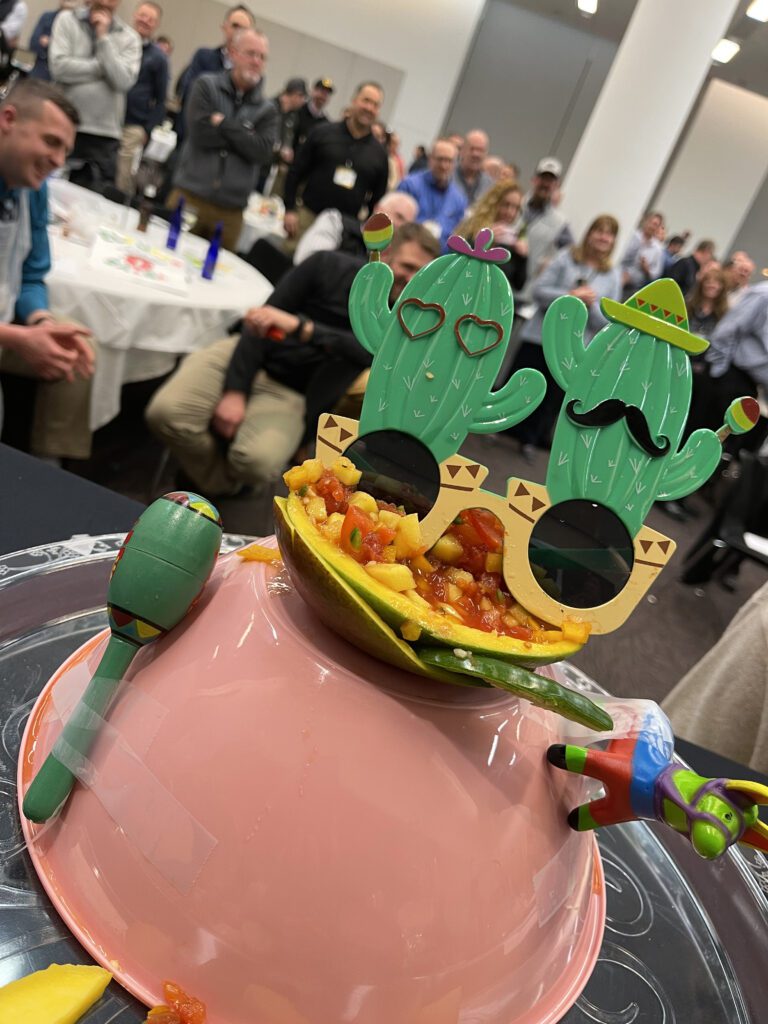 A festive salsa presentation featuring a bowl of fresh salsa garnished with playful cactus-shaped glasses, maracas, and a mini piñata, capturing the lively spirit of the Salsa Showdown team building event.