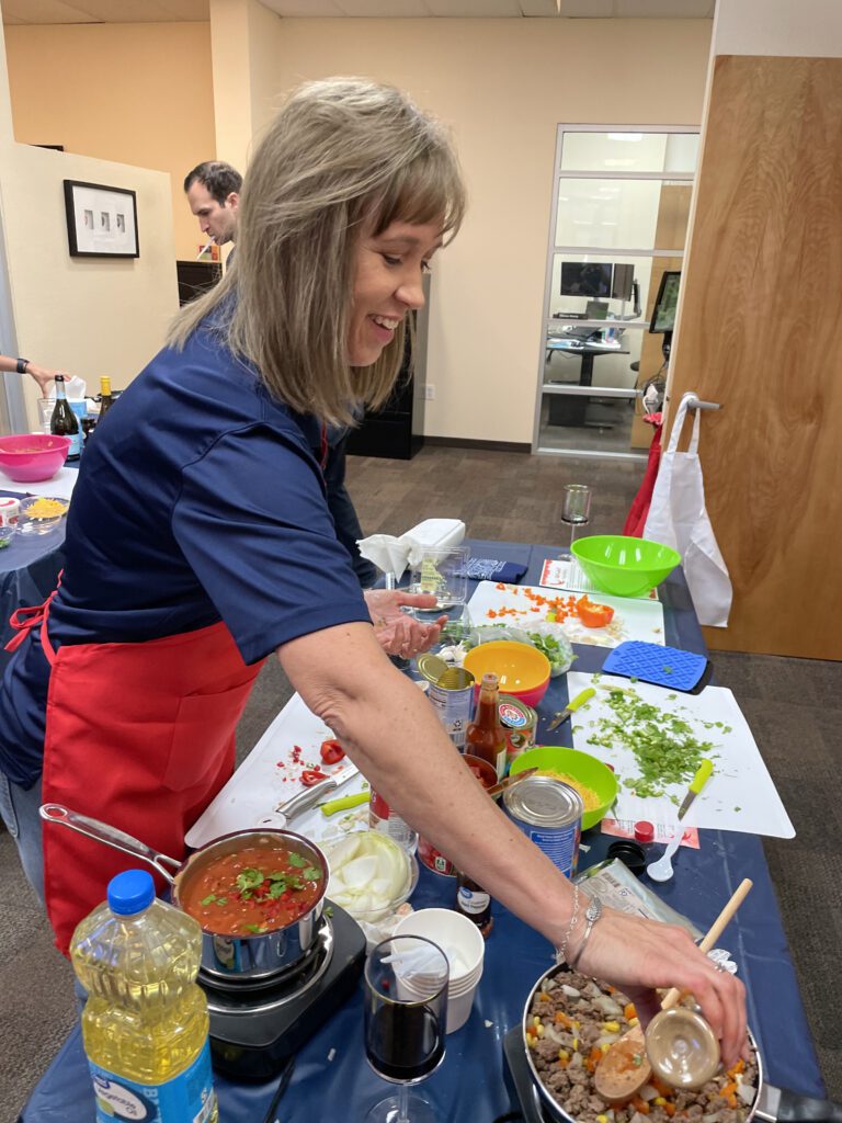 A woman is participating in the team building chili cook-off. She's wearing a red apron is smiling while cooking at a table filled with ingredients and kitchen tools. She is holding a small container to add seasoning to a pan filled with chopped vegetables and sausages. On the table, there are various ingredients, including cans, chopped onions, cilantro, a bottle of cooking oil, and a glass of red wine.