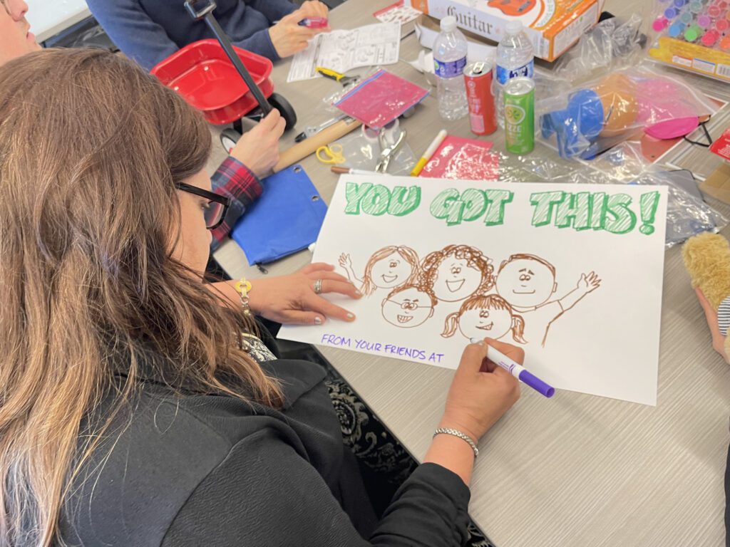 A person seated at a table working on a hand-drawn sign that reads 'You Got This!' The drawing includes smiling faces of people and the words 'From your friends at...' as part of the message. Surrounding the person are various arts and crafts supplies, this is part of a team building event.